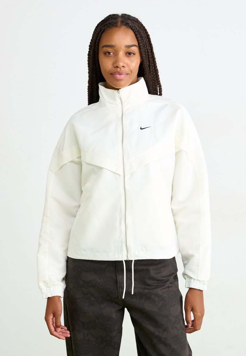 Woman with long braids wearing a white zip-up jacket with a small black Nike logo and dark patterned pants standing against a white background.