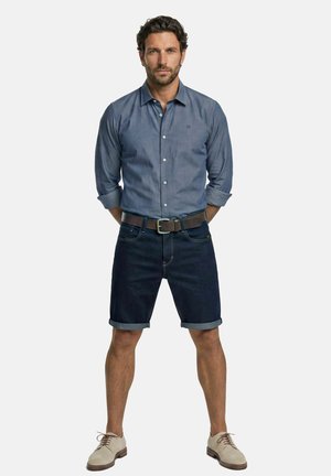 Mann mit dunklem Haar, der ein blaues Hemd mit Knöpfen, dunkle aufgerollte Jeansshorts, einen braunen Gürtel und beige Schnürschuhe trägt, steht mit den Händen hinter dem Rücken.