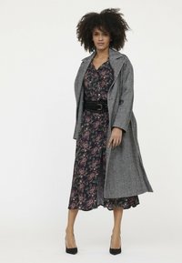 Grau gemusterter Haifischzahn-Trenchcoat über einem blumigen, dunklen Kleid mit einem taillierten Gürtel. Hochhackige schwarze Schuhe runden das Outfit ab.