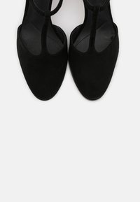 Chaussures à talons hauts en daim noir avec bout pointu et design à bride en T. Doublure intérieure lisse et dessus texturé, présentant un look minimaliste.