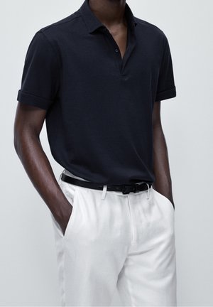 Navy polo shirt with a classic collar, short rolled sleeves, and a slim fit, paired with white linen trousers and a black belt.