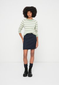 Jersey a rayas verdes y blancas con cuello redondo, combinado con una falda texturizada de color azul marino oscuro. Se lleva con botas negras con cordones y calcetines de punto.