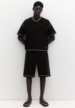 Jeune homme debout, les mains dans le dos, portant un t-shirt noir oversize et un short avec des coutures blanches et des sandales noires sur fond blanc.