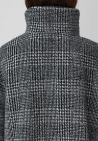 Szary i czarny płaszcz w kratę z wysokim kołnierzem. Tkanina ma miękką fakturę, z subtelnym wzorem i produktem z logiem na plecach.