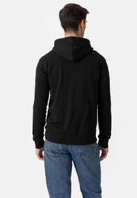 Črna jopa s fiksno kapuco, s gladko teksturo ter rebrastimi manšetami in spodnjim robom. Nošena z modrimi kavbojkami, prikazana od zadaj.