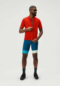 Rotes kurzärmeliges Radtrikot, blaue Shorts mit türkisfarbenen und weißen Akzenten sowie schwarze Radschuhe. Zubehör umfasst schwarze Sonnenbrillen.