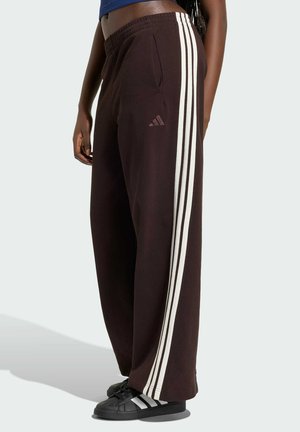 Osoba nosící černé tepláky Adidas s bílými postranními pruhy a černé tenisky s bílými pruhy, stojící bokem.