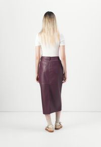 Burgunderfarget lær midi-skjørt med en tilpasset midje, baklommer og glatt tekstur. Modellen har på seg beige sokker og slip-on sandaler.