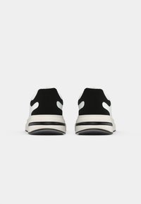 Scarpe sportive in bianco e nero con una tomaia liscia, un tallone nero testurizzato e una suola spessa bianca con una striscia nera.