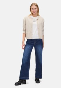 Beige texturerad cardigan med pärlknappar, bärs över en vit grafisk t-shirt och matchas med vida mörkdenim-jeans och svarta klackstövlar.