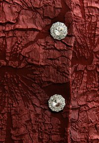 Tela burdeos con un patrón floral texturizado; presenta botones redondos de plata con una gema clara en el centro rodeada de piedras más pequeñas.