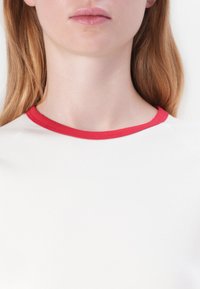 Biela tričko s okrúhlym výstrihom, ktoré má kontrastnú červenú rebrovanú obrubu, hladkú textúru a nemá viditeľné vzory ani logá.