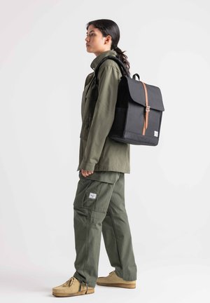 Black backpack with a flap closure, brown leather strap, rectangular shape, paired with an olive green jacket and cargo pants.
