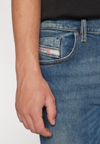 Jeans i blekblå denim med klassisk passform. Har en fickdetalj med en röd etikett och kontrasterande sömnad. Handen vilar nära fickan.