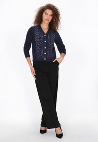Cárdigan de punto azul marino con cuello en V, que presenta patrones decorativos y botones plateados, combinado con pantalones de pierna ancha negros y tacones.