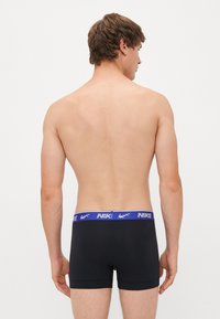 Hátulról látható férfi fekete boxer alsónadrágot visel, kék derékpánttal, amelyen fehér Nike logók találhatók, egyszínű háttérrel.