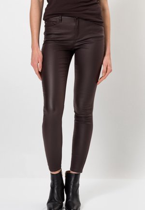 Slim fit dunkelbraune Lederhose, getragen mit schwarzen Stiefeletten, von der Taille bis zu den Füßen vor einem einfarbigen hellen Hintergrund gezeigt.