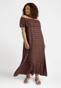 Abito maxi marrone con spalle scoperte, decorato con motivi diagonali dorati, in tessuto fluido e dalla vestibilità comoda. Indossato con sandali beige.
