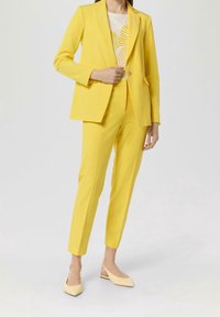 Costume jaune sur mesure avec blazer ajusté et pantalon fuselé, en tissu lisse. Associé à des ballerines jaune pâle et un haut léger.