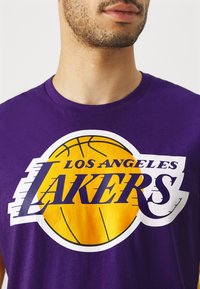 Vijolična bombažna majica s poudarjeno rumeno grafiko košarkarske žoge. Besedilo pravi "LOS ANGELES LAKERS" v krepki beli in vijolični pisavi.