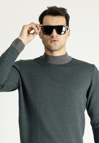 Maglione verde scuro con un motivo a maglia testurizzata e un colletto a coste grigio. Il modello indossa occhiali da sole neri, tenendoli con una mano.