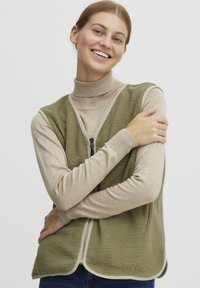 Zeleno flis telovnik z zadrgo spredaj, kremnim obvodom in mehkim videzom, nošen čez svetlo bež roll-neck pulover, na beli podlagi.