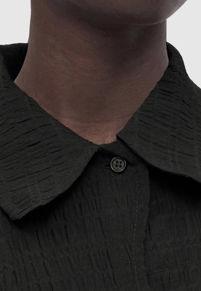 Σκούρος λαιμός και γραμμή σαγονιού που φοράει υφασμάτινο μαύρο πουκάμισο με γιακά, κουμπωμένο στο πάνω μέρος, σε απλό φόντο.