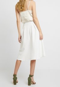 Nainen, jolla on päällä hihaton beige paita, valkoinen pleatattu midi-hame ja vihreät mokkasiinikorkonauhakengät, seisoo selin.