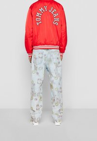 Giacca bomber rossa con la scritta "TOMMY JEANS" in lettere bianche, abbinata a jeans blu chiaro con motivi stampati colorati e una vestibilità rilassata.