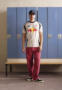 Bílý sportovní dres s černými akcenty a červeným logem, ve spojení s červenými kalhotami a černými botami. Stojí před modrými skříňkami.