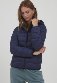 Námořnická modrá péřová bunda s kapucí, se zipovým zapínáním, prošívaným designem a elastickými manžetami. Nositá přes světle modrý top a šedé kalhoty.