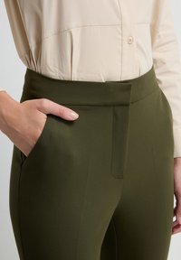 Olijfgroene broek met een gladde textuur, hoge taille en zijzak. Gecombineerd met een lichtbeige overhemd met knopen, met een getailleerde pasvorm.