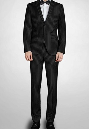 Homme portant un costume noir à fines rayures, une chemise blanche et un nœud papillon noir, debout devant un fond clair uni.