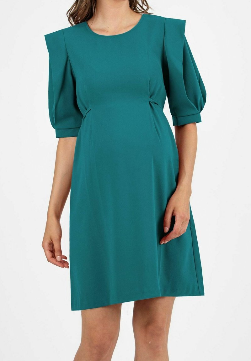 Robe bleu sarcelle longueur genou avec manches mi-longues bouffantes et épaules structurées portée par une personne avec les bras détendus sur les côtés.