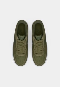 Zelené tenisky Nike Air Force 1 s hladkým povrchom, perforovanou špičkou a bielou gumovou podrážkou. Majú tonálne šnúrky a branding na jazyku.