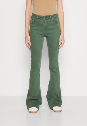 Jean flare verts en coton, avec fermeture à bouton, poches et détails de couture minimes. Portés avec des baskets blanches.