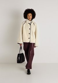 Witte fleecejas met hoge kraag, zwarte accenten en grote knopen, gecombineerd met bordeaux leren broeken en een zwarte tas, gedragen door een model.