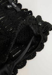 Tissu en crochet noir avec bords festonnés, fermeture par bouton et détail en ruban satin, affichant une étiquette de marque de vêtements.