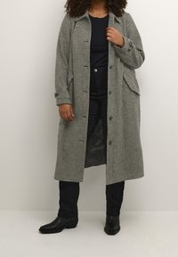 Kvinna i svarta stövlar, svarta byxor, svart skjorta och en lång svartvit houndstooth-mönstrad kappa som står mot en enkel vägg.