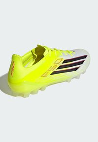 Φουτουριστικό παπούτσι ποδοσφαίρου σε νέον κίτρινο με λευκές λεπτομέρειες, υφασμένο επάνω μέρος, τρεις μαύρες ρίγες και ημιδιαφανές εξωτερικό πέλμα.