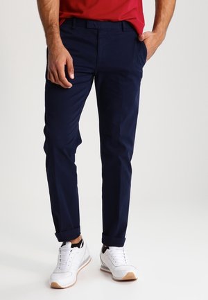 Homme portant un chino bleu marine, des baskets blanches et une chemise rouge, debout avec une main dans la poche devant un fond uni.