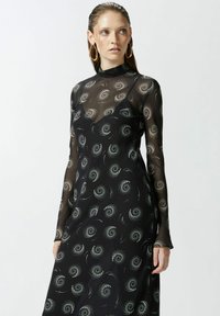 Crna haljina dugih rukava s prozirnim slojem, koja ima srebrne spiralne uzorke i visoki ovratnik. Glatka tkanina s lepršavim siluetom.