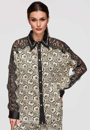 Femme portant un chemisier à motif floral noir et blanc avec manches en dentelle noire et un col noir clouté, posant avec une main sur la hanche.