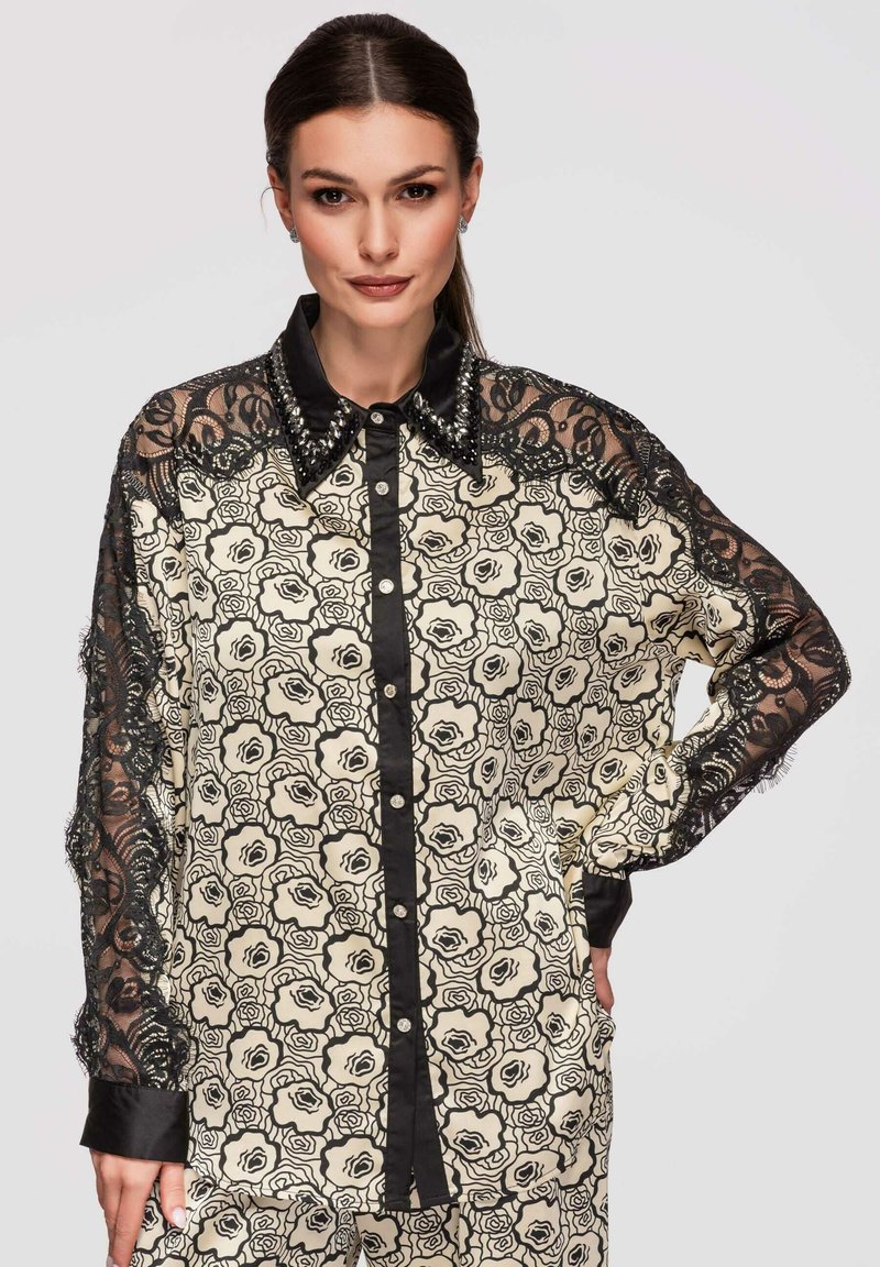 Femme portant un chemisier à motif floral noir et blanc avec manches en dentelle noire et un col noir clouté, posant avec une main sur la hanche.