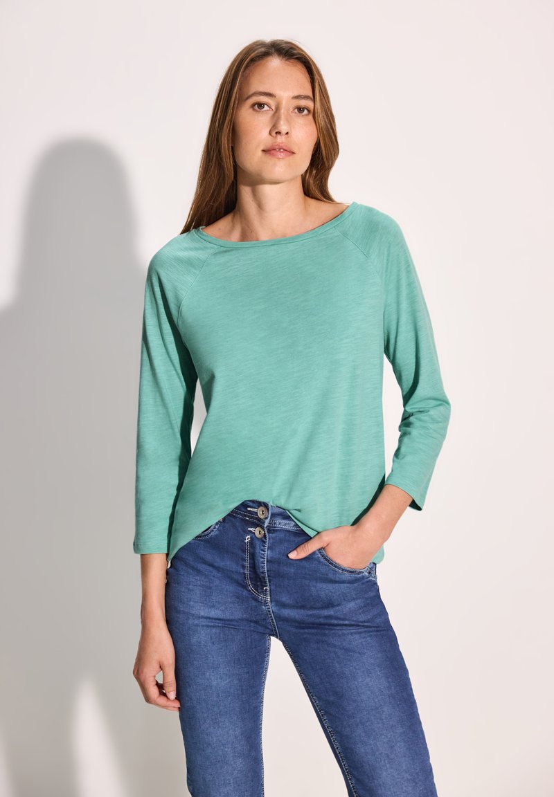 Ein mintgrünes, leichtes Langarmoberteil mit weitem Ausschnitt und abgerundetem Saum, kombiniert mit blauen Jeans, die mit einem Zweiknopfverschluss ausgestattet sind.