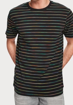 Uomo che indossa una maglietta nera a maniche corte con sottili strisce orizzontali arcobaleno, abbinata a pantaloni grigio chiaro, mani dietro la schiena.