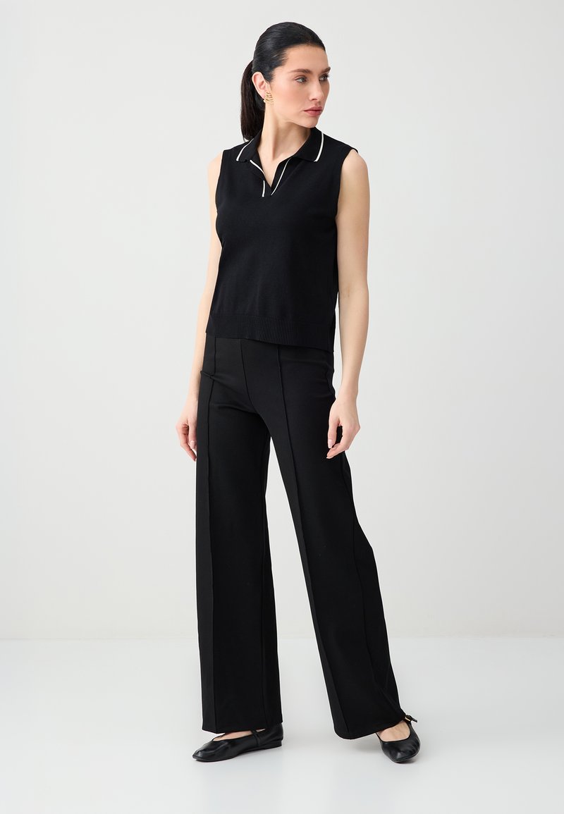 Femme debout portant un polo noir sans manches avec des bordures blanches, un pantalon noir à jambes larges et des ballerines noires, sur un fond clair uni.