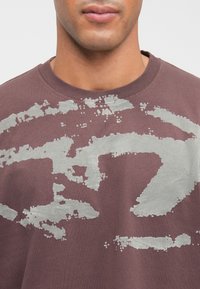 Muž v mauve tričku s okrúhlym výstrihom, na ktorom je veľký opotrebovaný abstraktný strieborný vzor cez hruď a plecia.