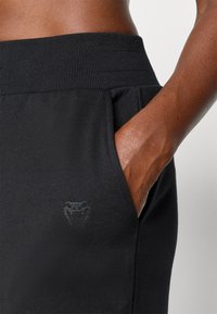 Zwarte sportbroek met een geribbelde tailleband, zijzakken en een subtiele ton-sur-ton logo nabij de zak. Het materiaal lijkt zacht en rekbaar te zijn.