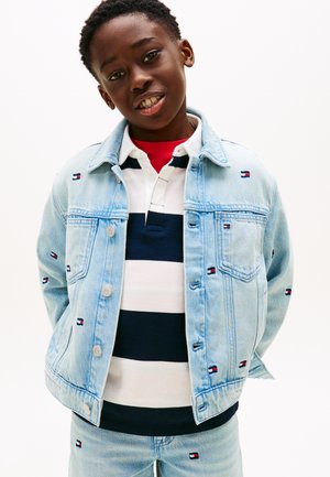 Ragazzo giovane sorridente, che indossa una giacca di jeans azzurro chiaro e jeans con piccoli loghi a bandiera, sopra una polo a righe bianche e blu navy.